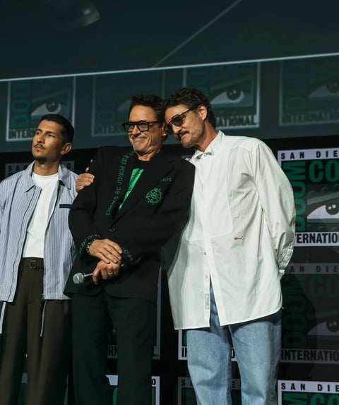 Three men standing on stage at San Diego Comic-Con, smiling and conversing with an eye-themed backdrop behind them