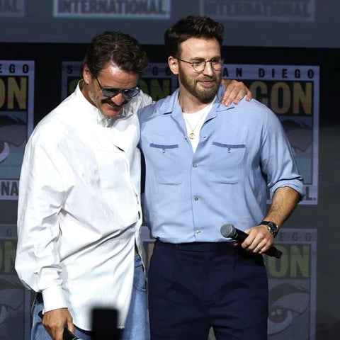 Two men at Comic-Con panel: one in white jacket and sunglasses, the other in light blue shirt holding a microphone